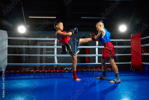 Confident two boys, kids, kickboxers, professional martial arts sportsmen performing kicks, fighting on ring at gym.