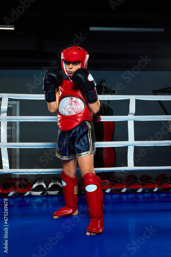 Child sportsman, boy in mouth guard dressed in uniform and protective gloves standing, posing on ring.