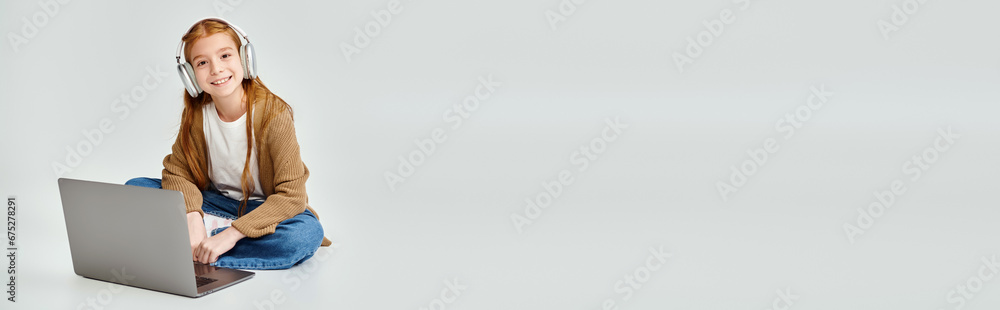 jolly preadolescent girl on floor with laptop and headset on gray backdrop, fashion concept, banner jolly preadolescent girl on floor with laptop and headset on gray backdrop, fashion concept, banner