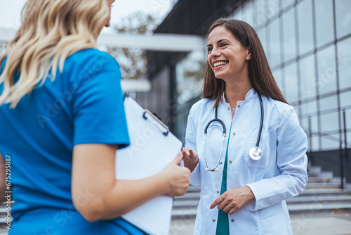 Two female doctor walk down an elevated walkway together. One wears a lab coat and the other wears scrubs. Female healthcare professionals walk and talk together photo
