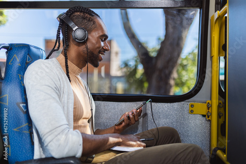 Jovem homem negro, usando fones de ouvido, ouvindo musica em seu smartphone, durante trajeto de onibus. photo