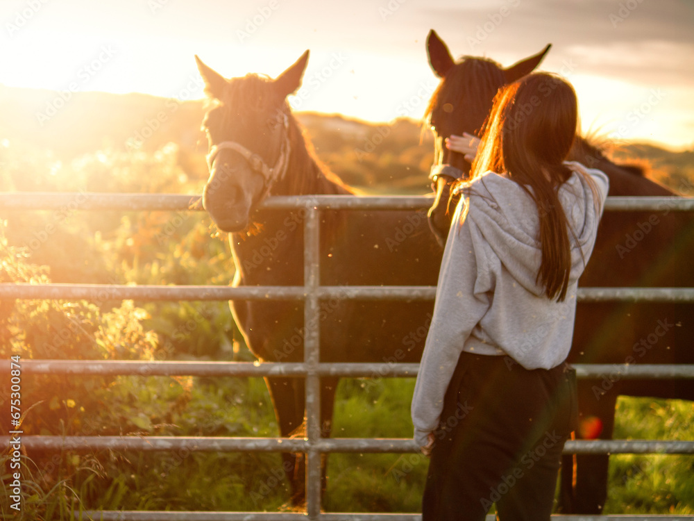 Obraz premium Teenager girl looking at dark horses by a metal gate to a field at stunning sunset. Warm sunshine glow. Selective focus. Light and airy soft look. Connection to nature concept. Rural area.