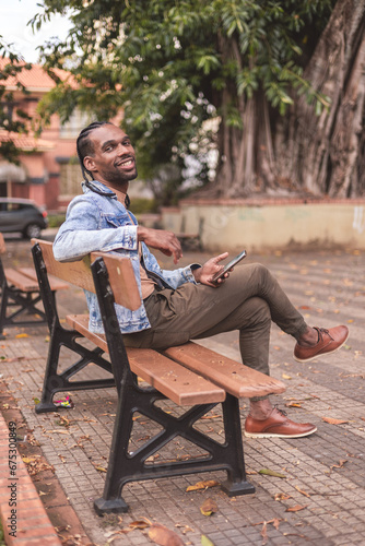 Young black man, sitting on the bench in the square, smiling, holding his smartphone.