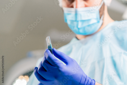 Close-up shot of a surgeon in a protective headdress and a medical mask holding scalpel in his hands and looking at it carefully photo