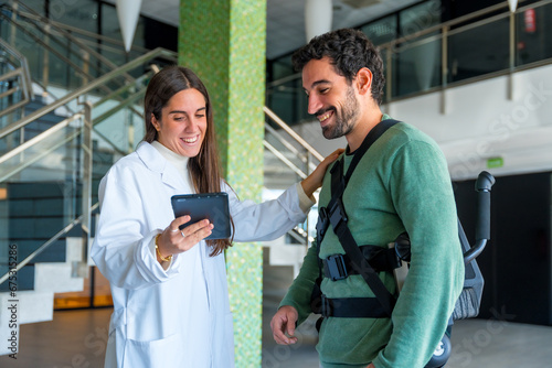Mechanical exoskeleton, female physiotherapist doctor talking and smiling with tablet with disabled person with robotic skeleton, physiotherapy in modern hospital, futuristic physiotherapy