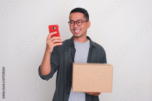 Adult Asian man smiling when looking to his phone while holding a box of package photo