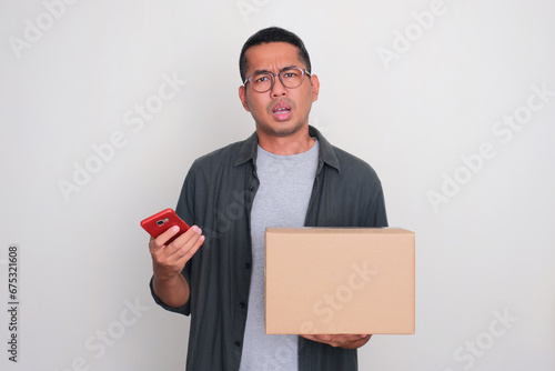 Adult Asian man showing confuse expression while holding phone and a box of package photo