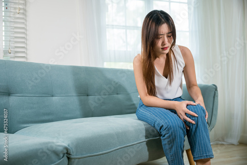 Asian woman massage hurts knee leg pain with painful face, young female sitting on sofa and feeling suffering knee pain in living room at home, Health bone problems self physiotherapy photo