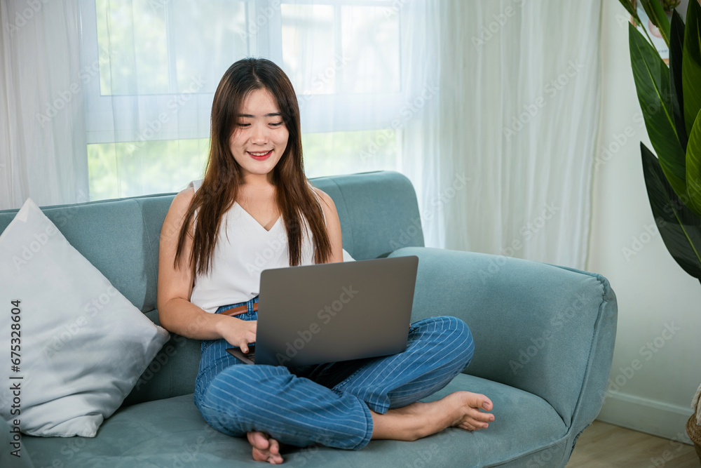 Asian young woman smiling sitting relaxing on sofa using laptop in living room at home, freelance working on computer project, Happy female typing email on notebook computer Asian young woman smiling sitting relaxing on sofa using laptop in living room at home, freelance working on computer project, Happy female typing email on notebook computer