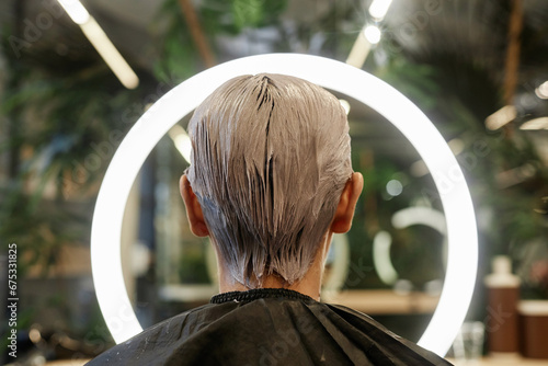 Back view of young man with bleached hair facing ring light in beauty salon and taking photos
