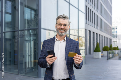 Portrait of a satisfied gray-haired businessman making an online purchase with a card in the phone in his hands, standing in front of a modern office, happy with the purchase, smiling at the camera.