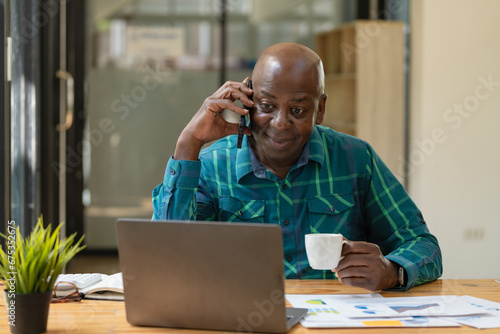 African American senior man with a coffee mug and using a smartphone while working with a laptop in an office.