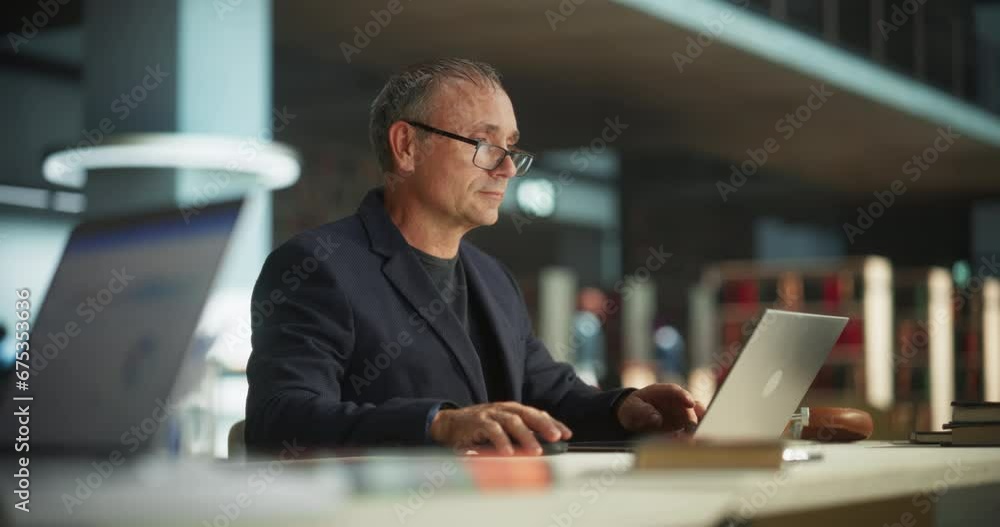 Knowledgeable Teacher Using Laptop Computer in a Library. Smart Middle Aged Male Educator Preparing Lectures for College Students, Checking Exams, Browsing and Researching Internet Articles Knowledgeable Teacher Using Laptop Computer in a Library. Smart Middle Aged Male Educator Preparing Lectures for College Students, Checking Exams, Browsing and Researching Internet Articles