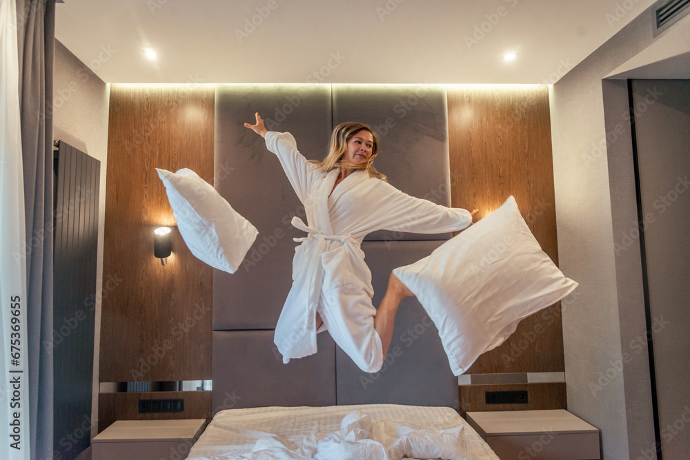 Woman having fun and jumping with bathrobe on bed in hotel room - obrazy, fototapety, plakaty 