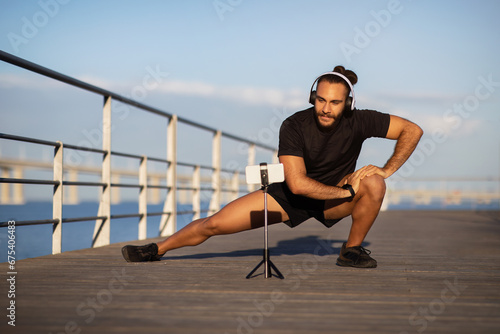 fitness guy in deep lunge, streaming exercise via smartphone outdoors © Prostock-studio