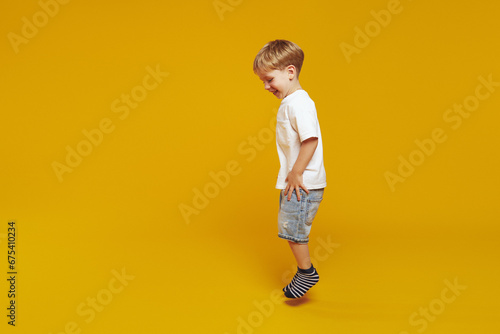 Horizontal image of side view full body stylish little kid boy in white t-shirt and striped socks, jumping while smiling against yellow background.