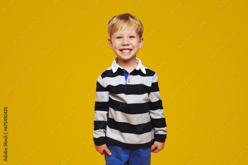 Excited handsome child with blonde hair, wearing striped shirt, smiling broadly for camera while standing against yellow background. Children studio portrait. Excited handsome child with blonde hair, wearing striped shirt, smiling broadly for camera while standing against yellow background. Children studio portrait.