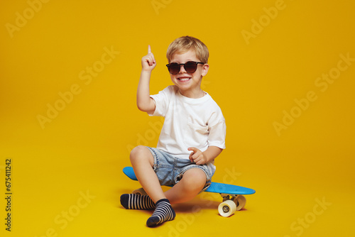 Adorable little boy in white tshirt and sunglasses, sitting on modern skateboard while looking at camera and pointing up, against yellow background.