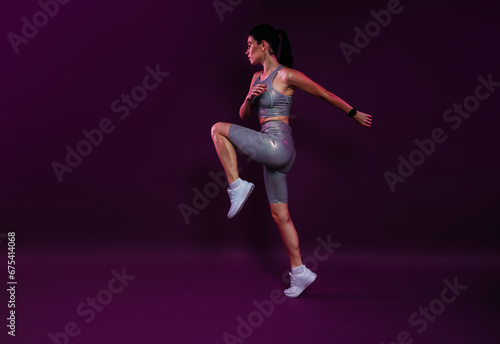 Full length of a young slim jogger practicing over a magenta backdrop. Female in silver fitness attire jumping.