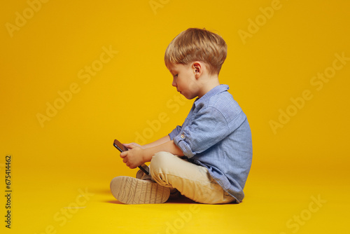 Side view of focused little boy sitting on studio floor with crossed legs while playing addictive games on smartphone, isolated over yellow background. Gadget addiction. photo