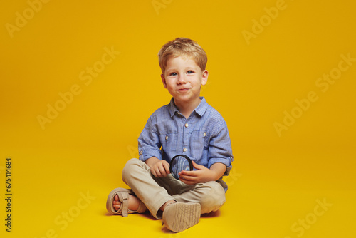 Little blonde haired boy in trendy blue shirt holding magnifier and looking at camera while sitting on studio floor with crossed legs, isolated over yellow background.