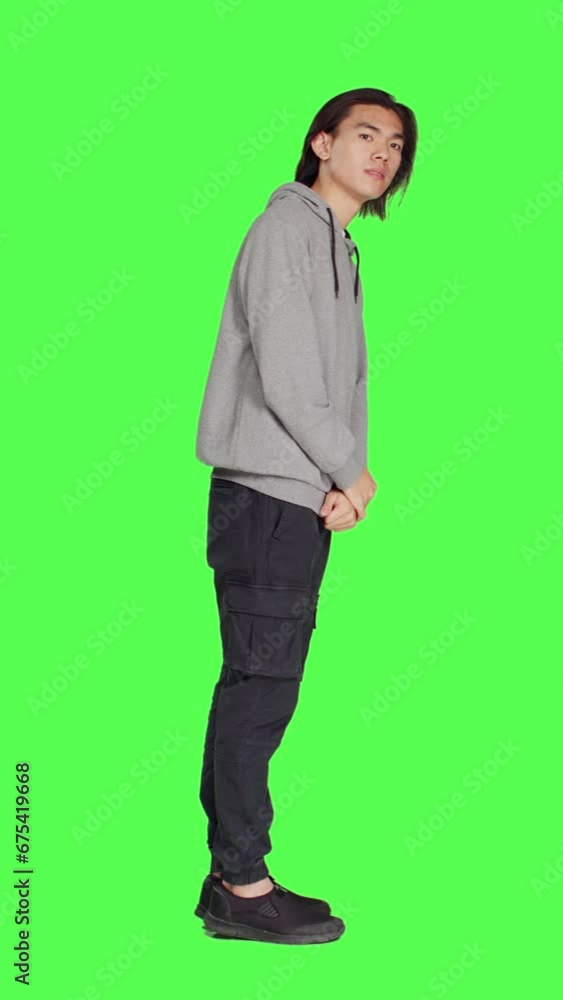 Profile in studio, young guy poses against greenscreen, looking at wristwatch and appearing distressed. Unhappy and irritated asian person is impatiently awaiting for stuff on camera. Profile in studio, young guy poses against greenscreen, looking at wristwatch and appearing distressed. Unhappy and irritated asian person is impatiently awaiting for stuff on camera.