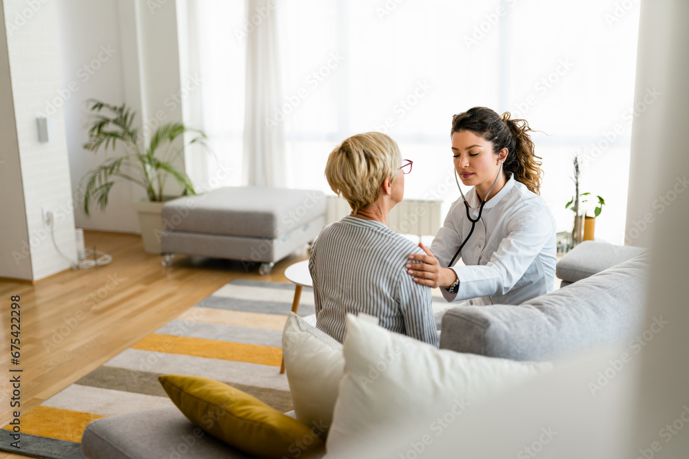 Attractive young female doctor checking senior patient with stethoscope at home visit while sitting on sofa in living room.