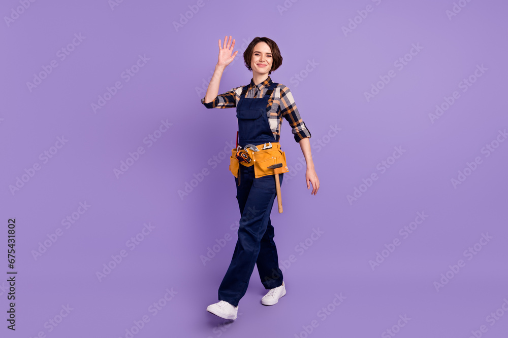 Full length profile side photo of young woman happy smile repairer go walk step wave hello isolated over violet color background Full length profile side photo of young woman happy smile repairer go walk step wave hello isolated over violet color background