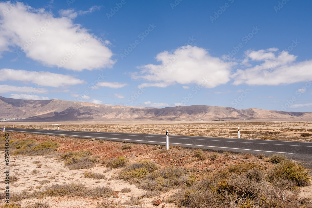 Desert landscape of white sand and desert shrubs. Asphalt road, mountains in the background. Sky with big white clouds. Lanzarote, Canary Islands, Spain.