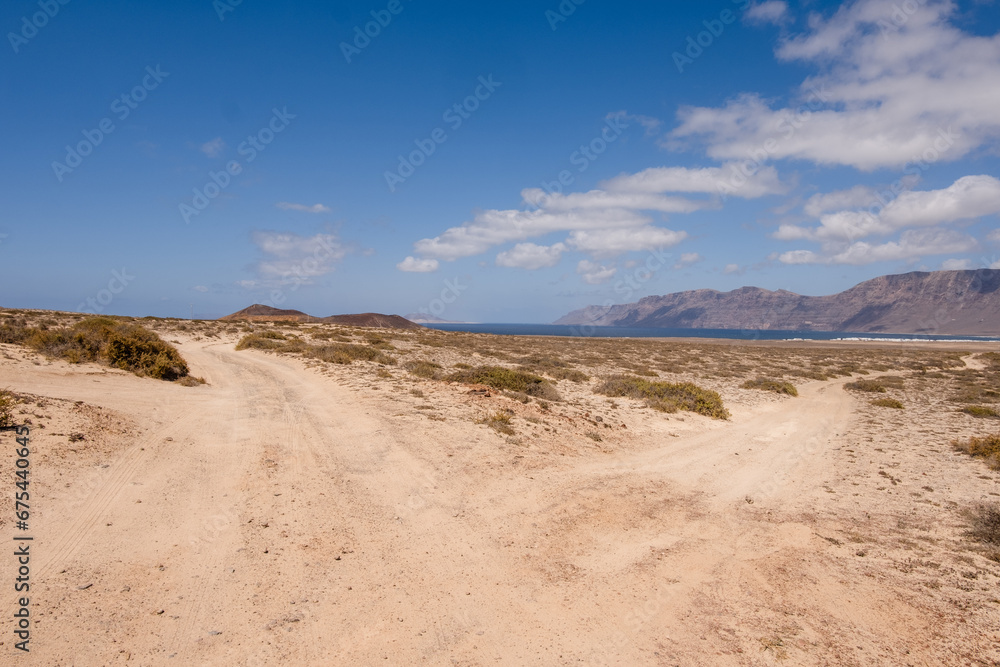 Desert landscape of white sand and desert bushes. Ocean and Famara cliff in the background. Dirt track. Sky with big white clouds. Lanzarote, Canary Islands, Spain.