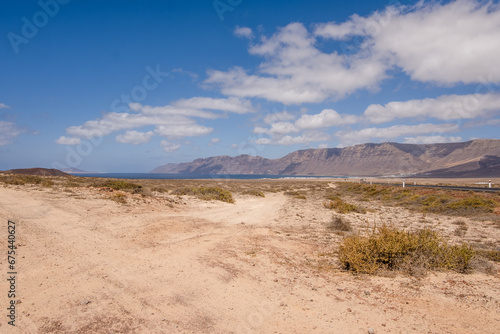 Desert landscape of white sand and desert bushes. Ocean and Famara cliff in the background. Dirt track. Sky with big white clouds. Lanzarote, Canary Islands, Spain.