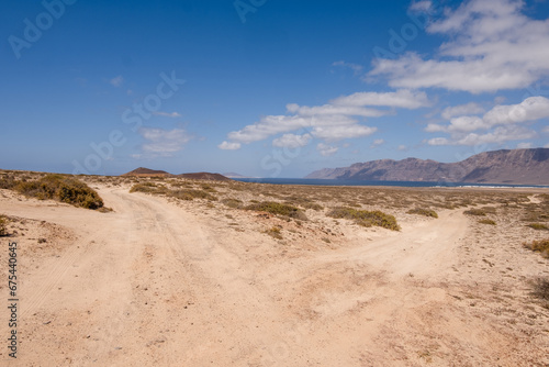 Desert landscape of white sand and desert bushes. Ocean and Famara cliff in the background. Dirt track. Sky with big white clouds. Lanzarote  Canary Islands  Spain.