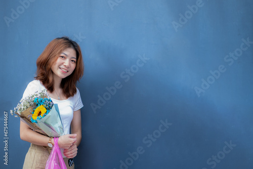 A white teenage girl smiles happily next to a blue wall and holds his favorite flower, waiting for someone.