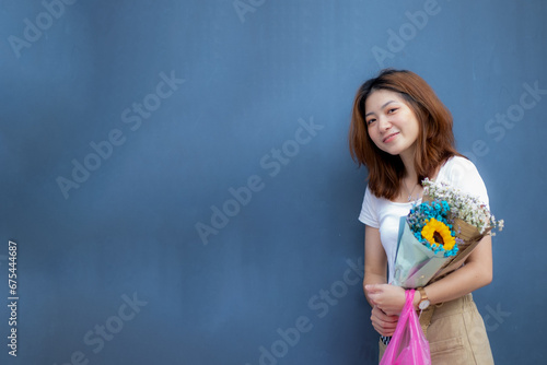 A white teenage girl smiles happily next to a blue wall and holds his favorite flower, waiting for someone.