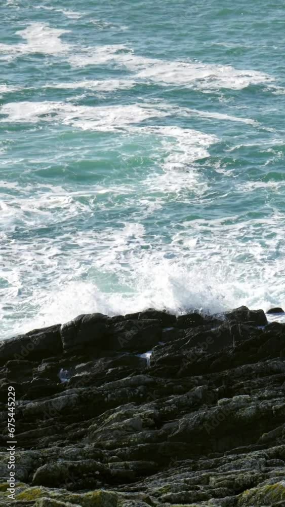 A rocky coastline characterized by its dark and jagged rocks, unyielding against the relentless assault of ocean waves. The deep blue-green sea, crashing waves, white foam. Vertical video. A rocky coastline characterized by its dark and jagged rocks, unyielding against the relentless assault of ocean waves. The deep blue-green sea, crashing waves, white foam. Vertical video.