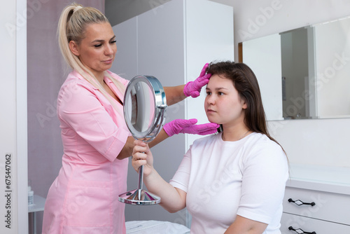 Beautician Prepares Plus Size Patient To Facial Dermal Filler, Beauty Procedure. Young Pretty Woman Looks At Mirror In Spa Salon. Female Receiving Plastic Surgery Injection On Face. Horizontal Plane
