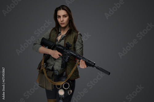 Middle Eastern-looking woman dressed in survivalist clothing posing with a rifle against a gray background, portraying strength and resilience © Fxquadro