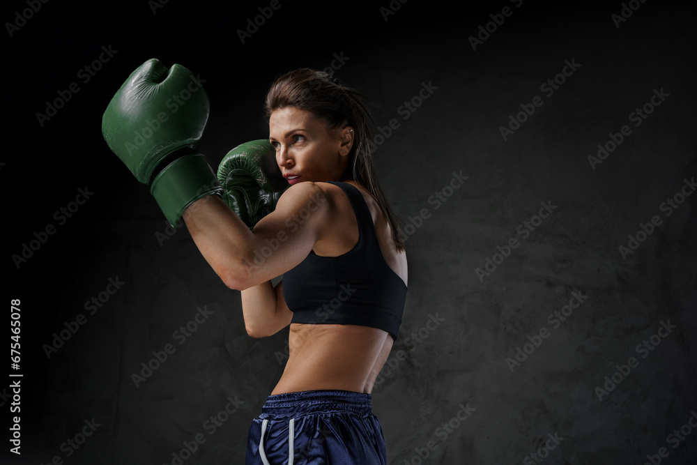 Athletic female fighter in sports bra, shorts, and boxing gloves showcases striking techniques against a dark backdrop