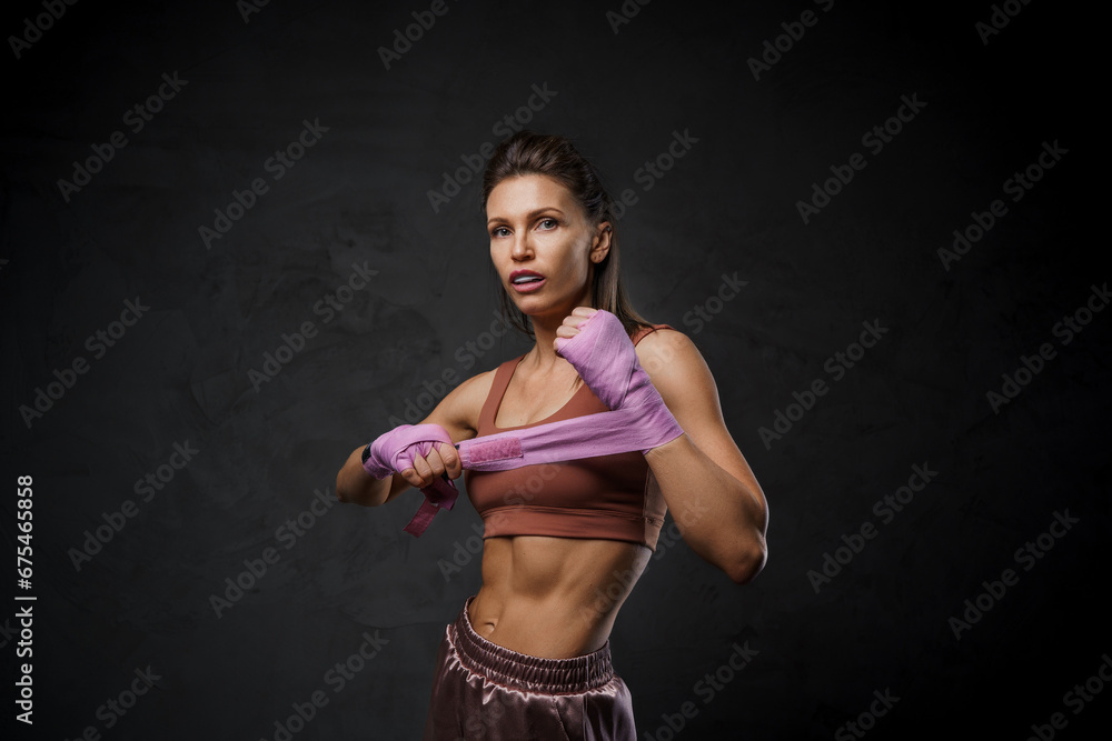 Obraz premium Athletic woman fighter in sports bra and shorts wraps her fists with boxing bandages against a dark backdrop