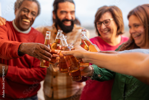 Multiracial friends toasting beer bottle in brewery pub garden - Happy different age people enjoying happy hour sitting in bar restaurant - Friendship concept