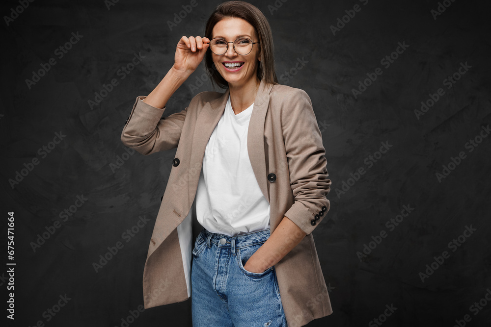 Joyful, stylishly dressed woman in glasses, tee, and blazer poses against a dark background