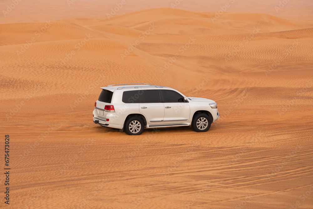 White SUV truck driving on a sand dune near Dubai, UAE at sunset hour, motion blur on the wheels. Extreme sports, adventure and travel concept. Wide angle shot.