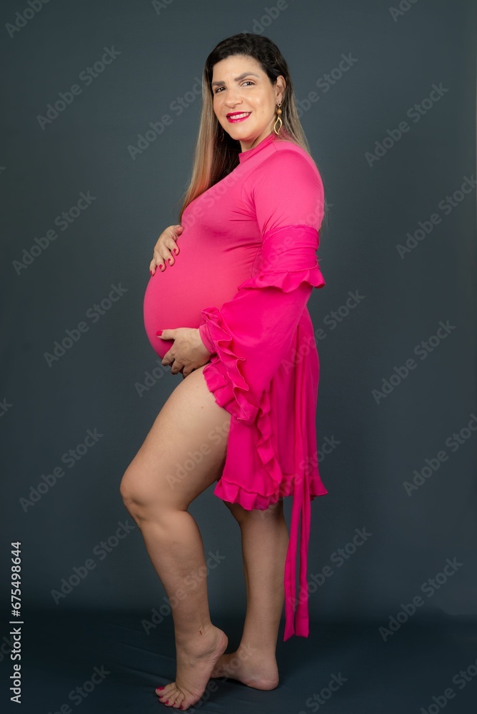 Obraz premium Young cheerful pregnant female in a pink dress smiles happily and contentedly posing in the studio