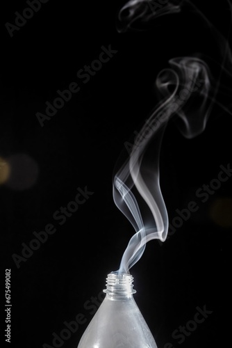 Plastic bottle with its lid off emits a plume of smoke into the air on the dark background