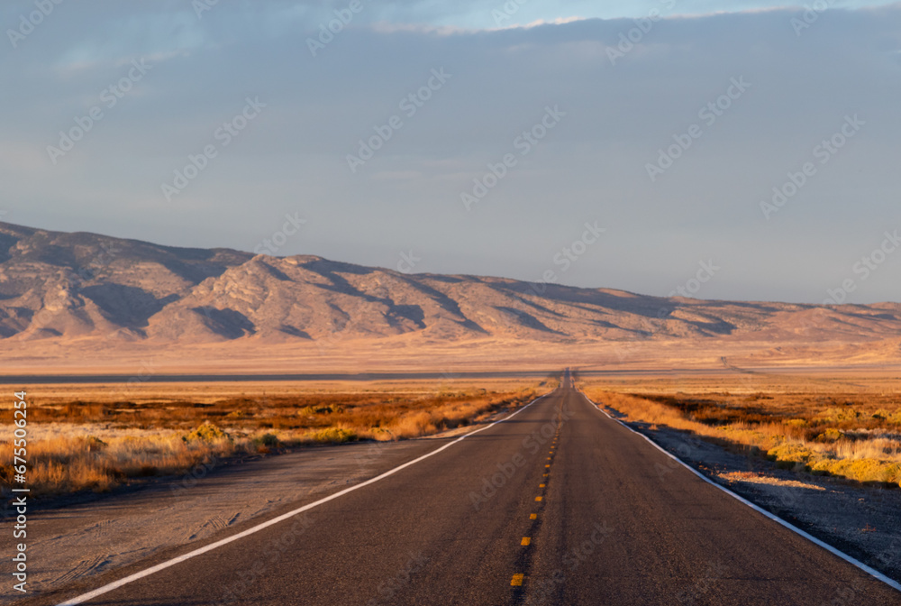sunset in the desert, image shows the beautiful golden sunset glow in the Nevada desert with some clouds, a empty long asphalt road and a mountain view in the background, taken october 2023