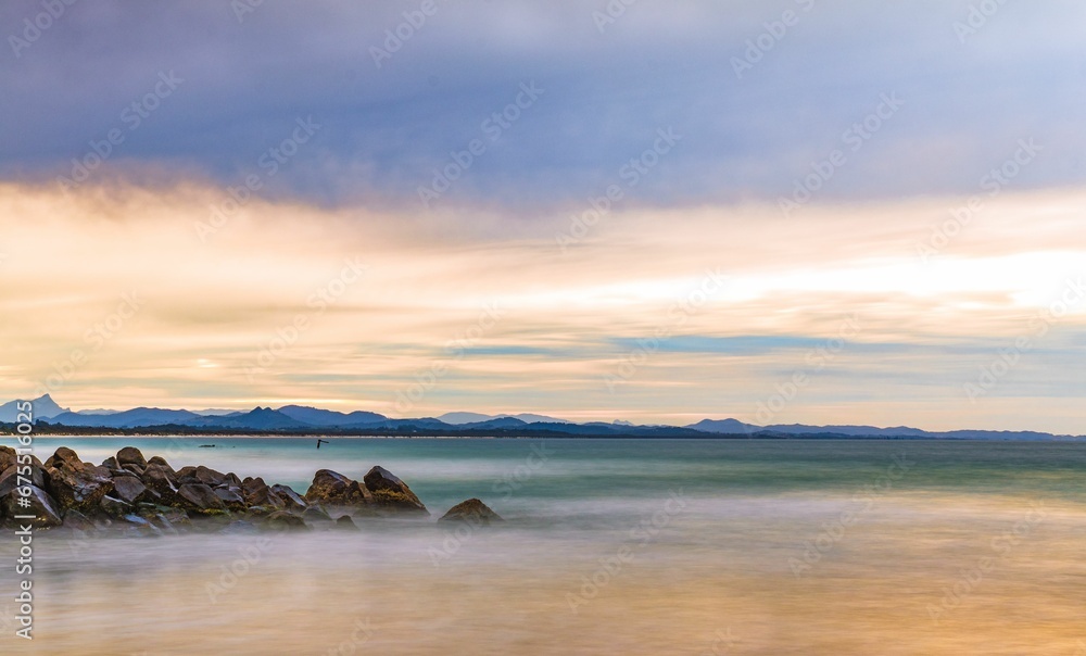 Long exposure shot of sandy beach shoreline against a backdrop of rocky outcrops and a cloudy sky. Long exposure shot of sandy beach shoreline against a backdrop of rocky outcrops and a cloudy sky.