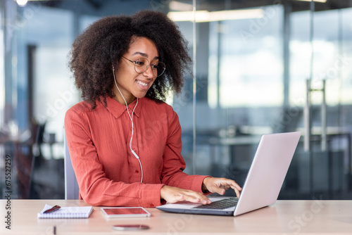 Young beautiful woman inside the office works with a laptop, a businesswoman in headphones listens music, podcasts, audio books and training course. Worker smiling , with curly hair and red shirt photo