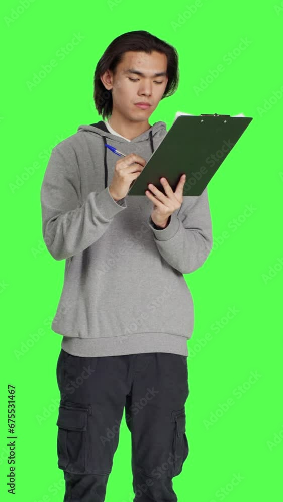 Front view of asian guy reads information on files, thinking about annotations on clipboard papers or taking notes for completion. Young man writing on forms, standing against greenscreen backdrop. Front view of asian guy reads information on files, thinking about annotations on clipboard papers or taking notes for completion. Young man writing on forms, standing against greenscreen backdrop.