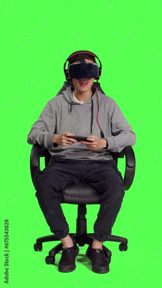 Front view of person rests on chair over greenscreen backdrop and performs video games using virtual reality eyewear and mobile device, takes part in online multiplayer contest. Asian guy loving Front view of person rests on chair over greenscreen backdrop and performs video games using virtual reality eyewear and mobile device, takes part in online multiplayer contest. Asian guy loving