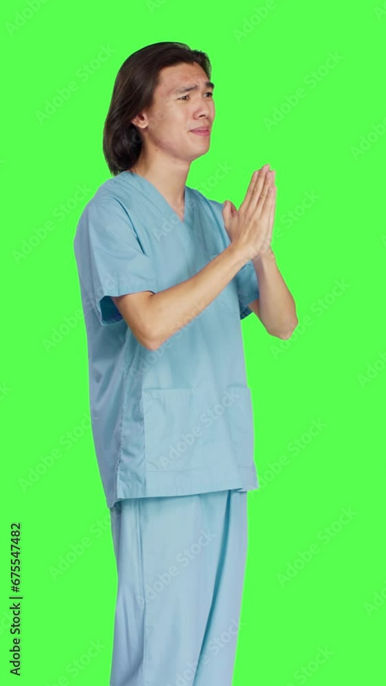 Side view of medical assistant praying to Jesus in studio, holding hands in a prayer while he stands against greenscreen background. Young male nurse with spiritual belief, showing hope. Side view of medical assistant praying to Jesus in studio, holding hands in a prayer while he stands against greenscreen background. Young male nurse with spiritual belief, showing hope.